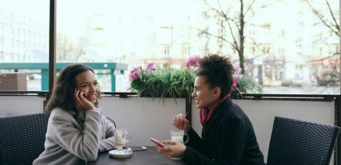 Two women friends at an outdoor cafe table, one talking on her phone while the other looks at her smartphone, a picture of friends looking out for each other when dating