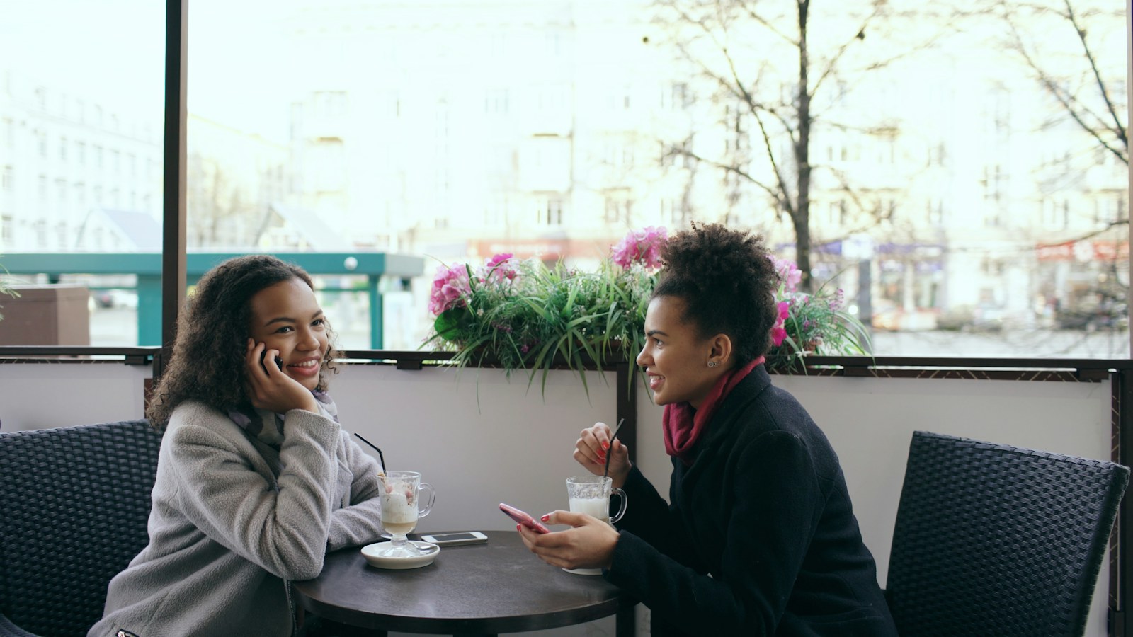 Two women friends at an outdoor cafe table, one talking on her phone while the other looks at her smartphone, a picture of friends looking out for each other when dating