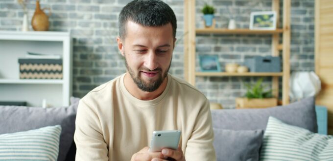 A man sitting on a couch looking at his smartphone, waiting on a reply to a text message